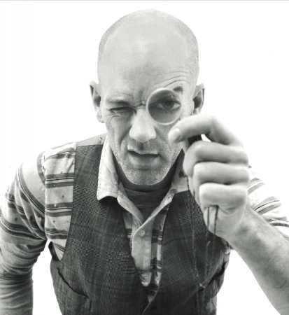 Black and white portrait of a mature adult male winking while looking through a monocle, wearing a vest and collared shirt.