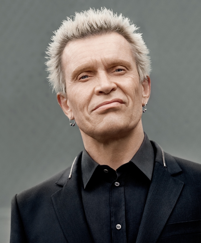 Portrait of musician Billy Idol, featuring his distinctive platinum blonde spiky hair, dressed in a black suit against a neutral gray backdrop.