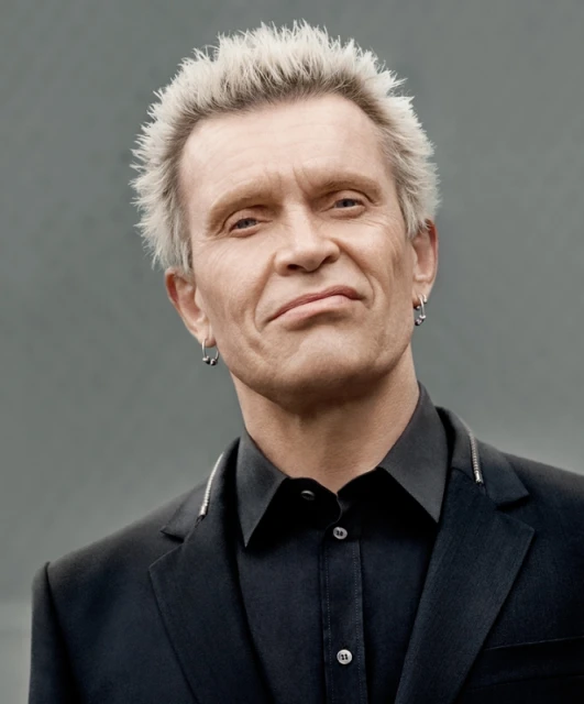 Portrait of musician Billy Idol, featuring his distinctive platinum blonde spiky hair, dressed in a black suit against a neutral gray backdrop.