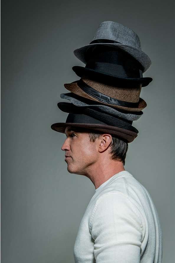 Man in profile wearing a tall stack of fedora hats in shades of brown, grey, and black against a plain grey background. Fashion still life.