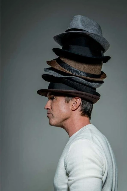 Man in profile wearing a tall stack of fedora hats in shades of brown, grey, and black against a plain grey background. Fashion still life.