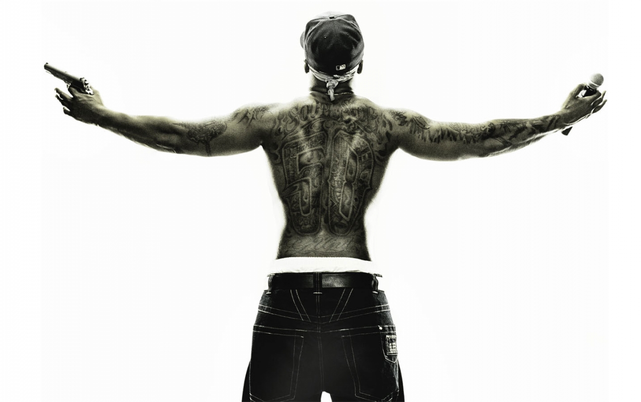 Back view of a shirtless man with tattoos holding a gun and microphone, wearing a baseball cap and jeans, on a bright white background.