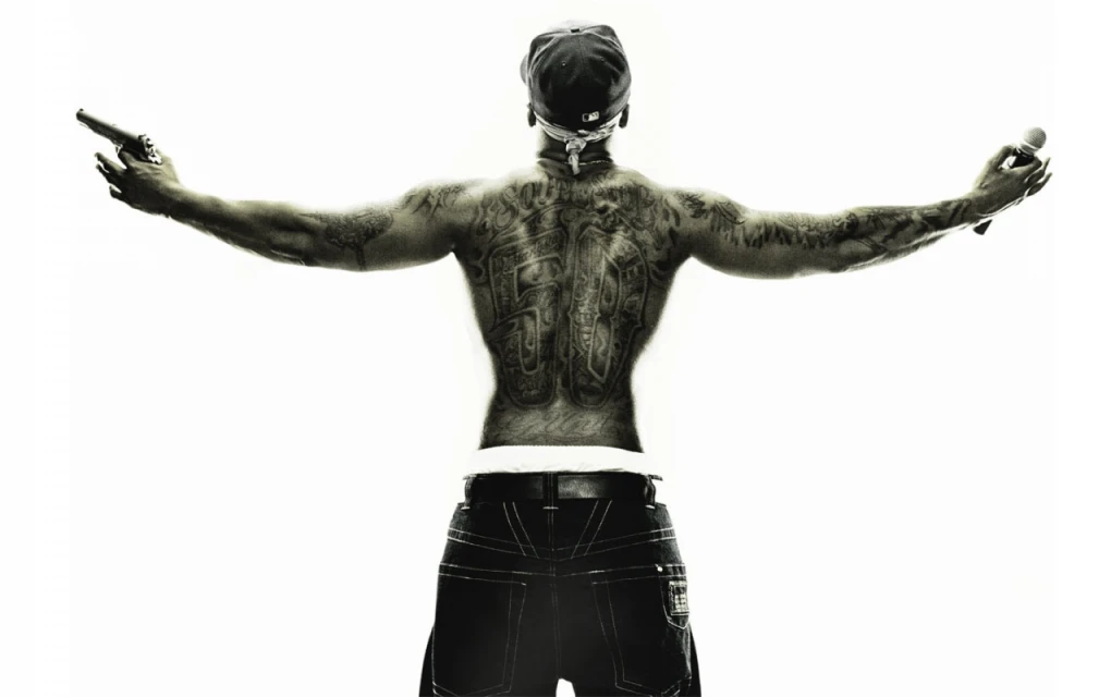 Back view of a shirtless man with tattoos holding a gun and microphone, wearing a baseball cap and jeans, on a bright white background.