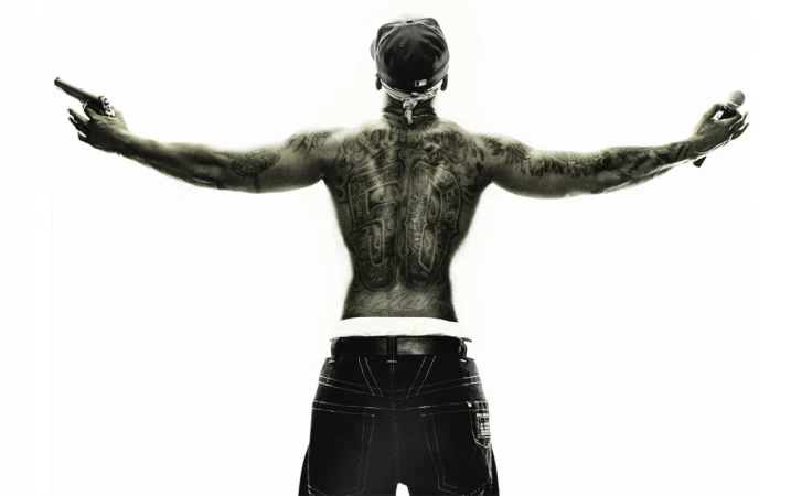 Back view of a shirtless man with tattoos holding a gun and microphone, wearing a baseball cap and jeans, on a bright white background.