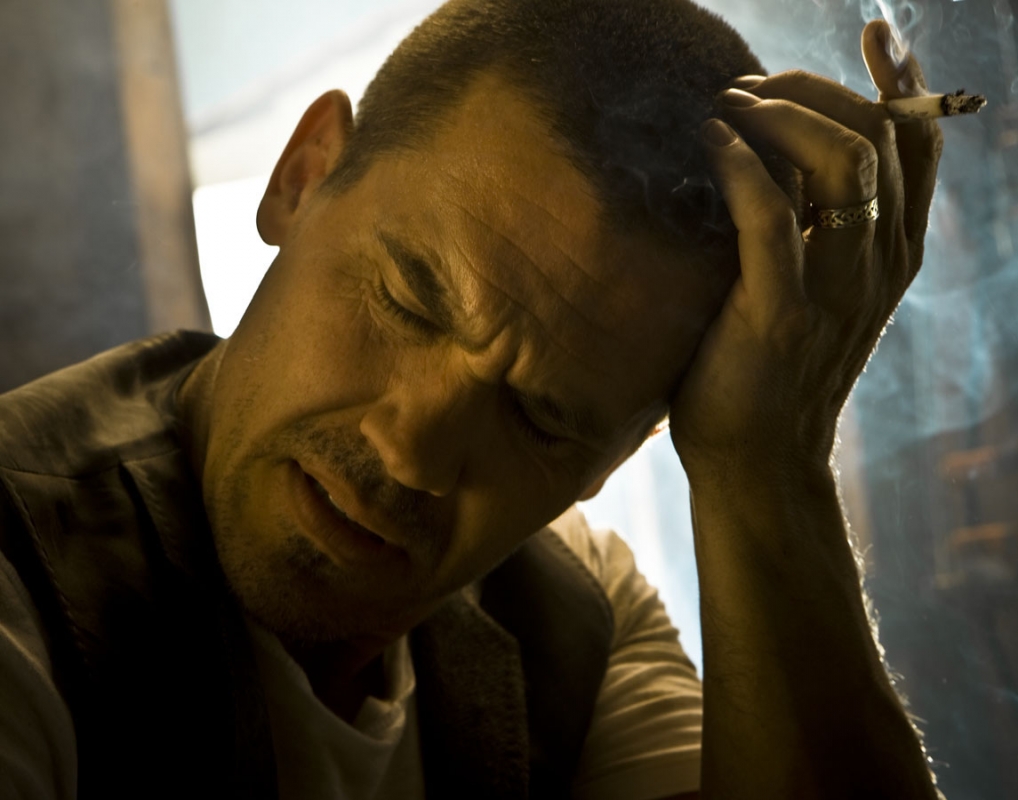 Man with headache smoking cigarette, close up. Adult male with closed eyes, holding head with hand, wearing vest and t-shirt. Indoors with smoke.