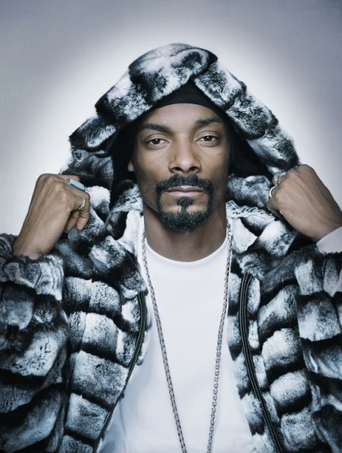 Portrait of Snoop Dogg in a fur-trimmed hooded coat, silver chains, and rings against a gradient gray background in a photo studio.