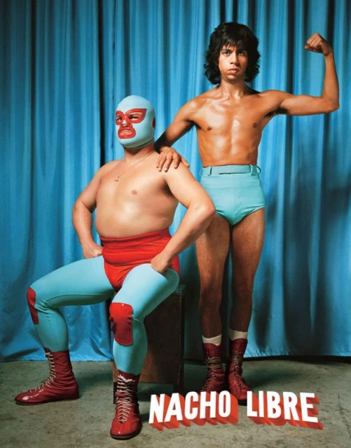 Two shirtless wrestlers pose in front of a blue curtain; one wears a blue and red mask, and the other is flexing an arm.