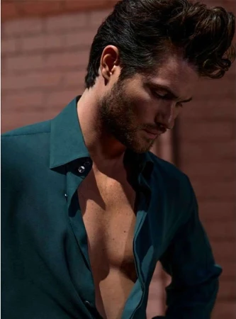 Close-up portrait of a man with dark hair and a beard wearing a dark green button-down shirt outdoors.