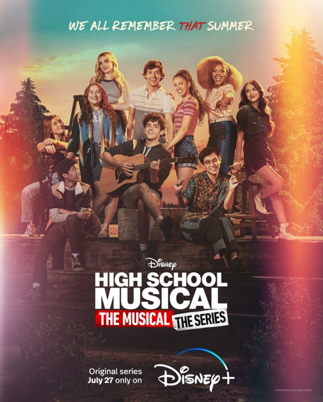 Group of young adult cast members from High School Musical: The Musical: The Series sit on a wooden structure outdoors.