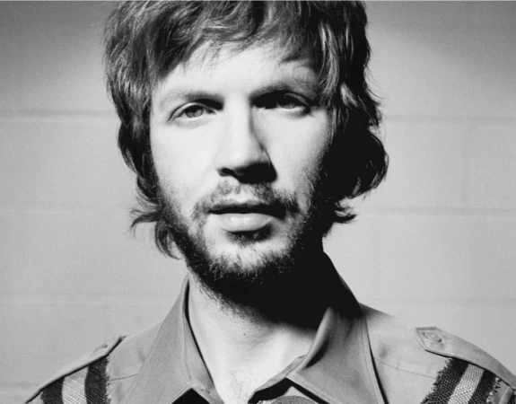 Portrait of Beck with shaggy hair and short beard wearing a collared shirt against a light tiled wall, photographed by Sheryl Nields.