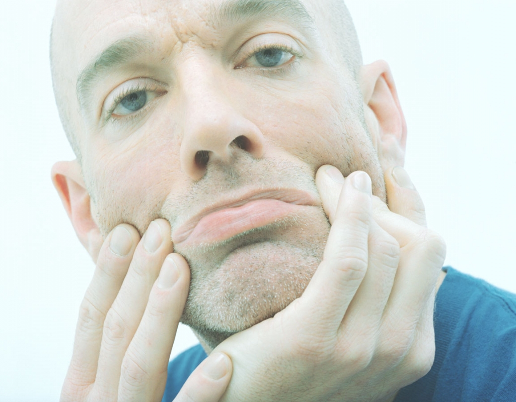 Close-up portrait of a pensive, bald adult male with blue eyes resting his face in his hands against a light, neutral background.