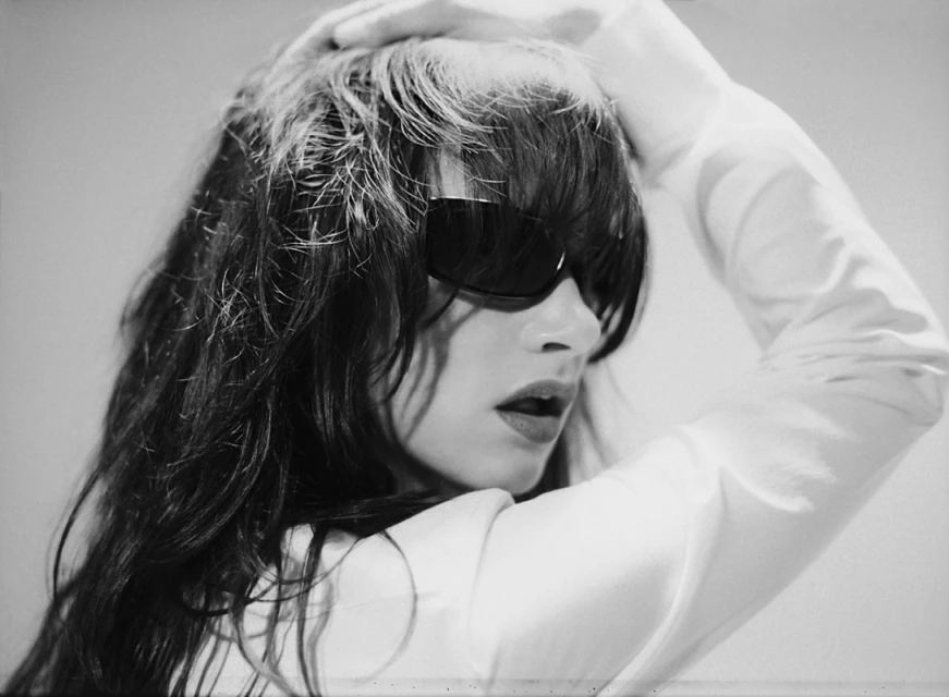 Black and white portrait of a woman with dark hair, sunglasses, and a white shirt posing with a hand on her head in a studio.