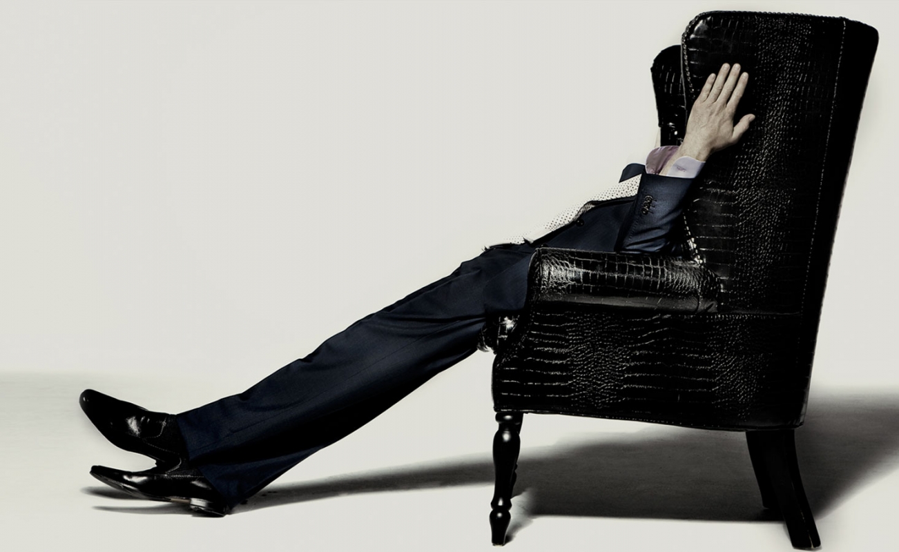 Man in dark suit reclines in a black wingback chair, legs stretched out, in a studio setting against a pale background.