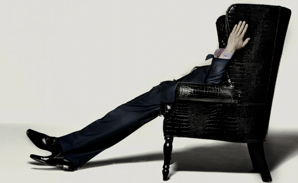 Man in dark suit reclines in a black wingback chair, legs stretched out, in a studio setting against a pale background.
