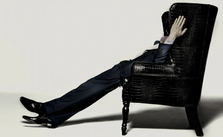 Man in dark suit reclines in a black wingback chair, legs stretched out, in a studio setting against a pale background.
