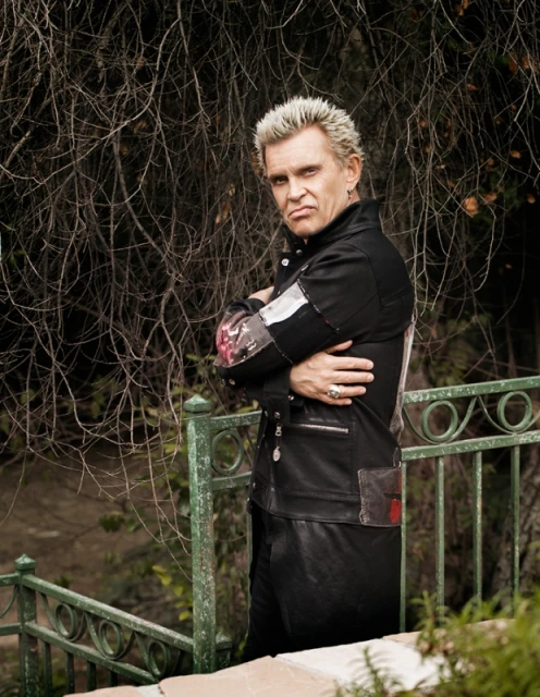 Billy Idol poses in a black leather jacket against an outdoor backdrop of trees and vegetation, with a metallic green stair rail.
