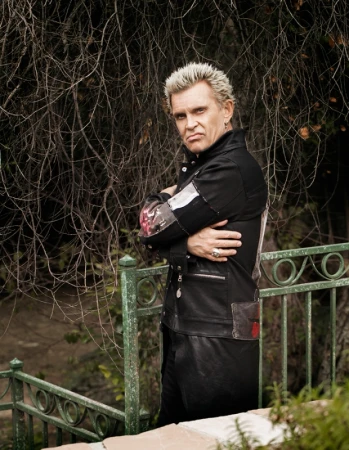Billy Idol poses in a black leather jacket against an outdoor backdrop of trees and vegetation, with a metallic green stair rail.