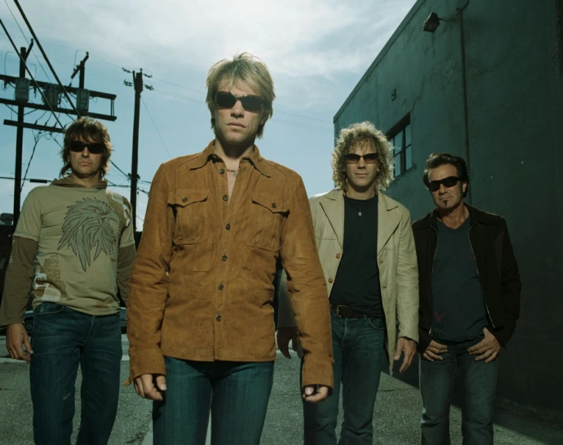 Four adult males stand outdoors on a city street wearing sunglasses. The central male is wearing a brown shirt and the others wear jackets.