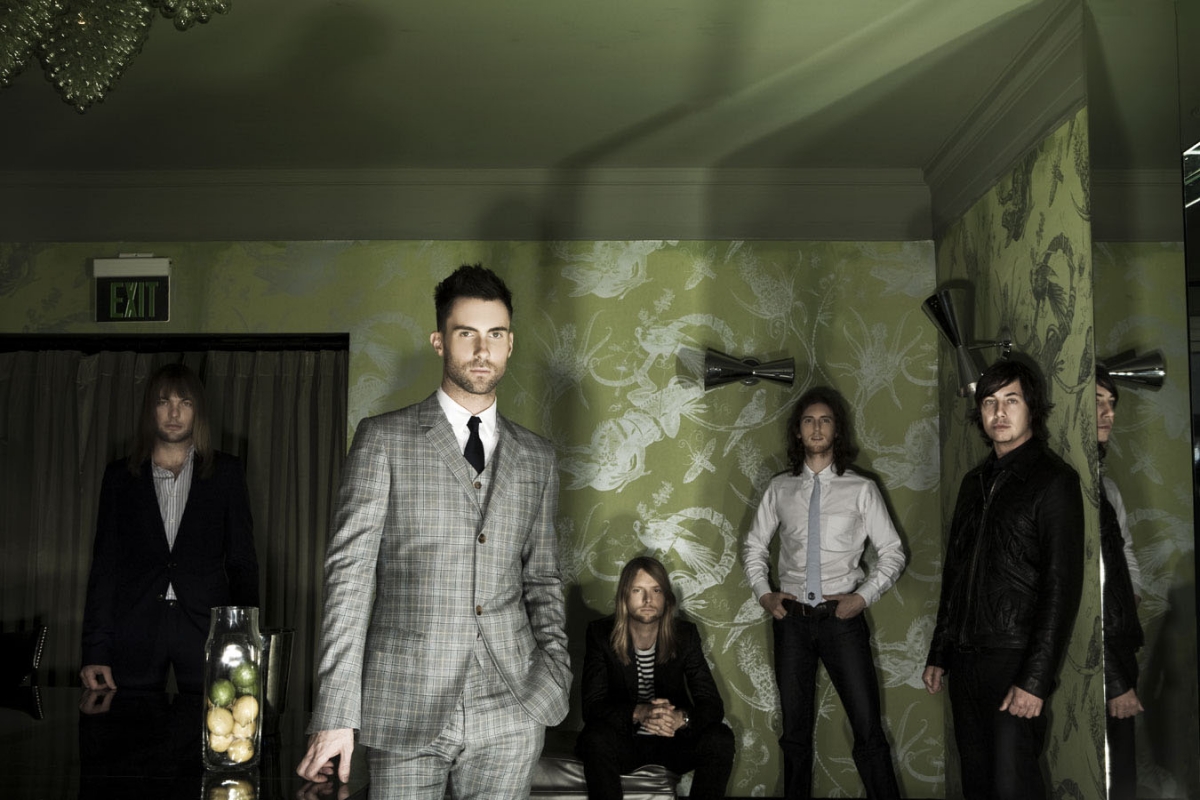 Band members pose indoors in suits and ties with lime green floral wallpaper, moody lighting, and modern chandelier.