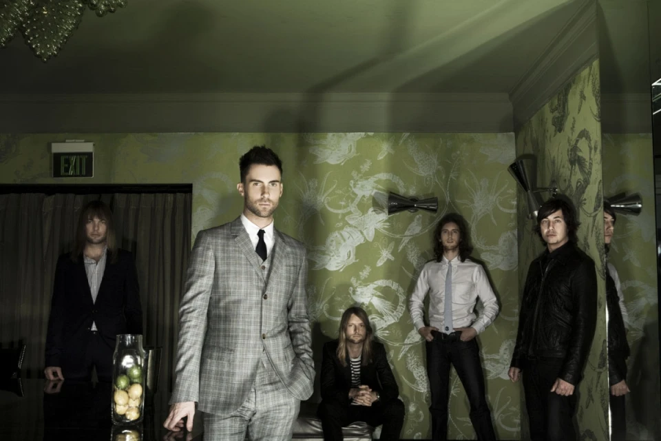 Band members pose indoors in suits and ties with lime green floral wallpaper, moody lighting, and modern chandelier.