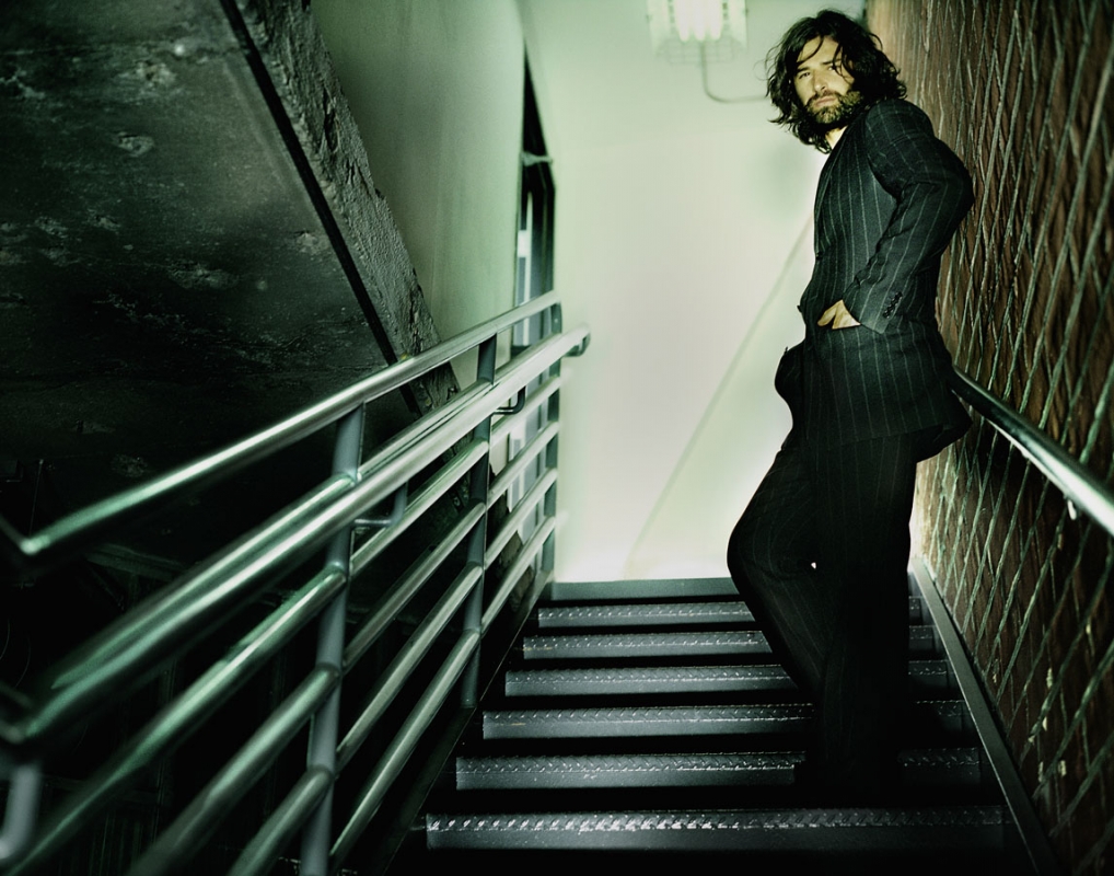 Man with long dark hair and beard, wearing a pinstripe suit, poses on metal stairs indoors with walls of concrete and brick.