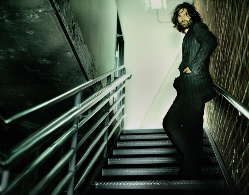 Man with long dark hair and beard, wearing a pinstripe suit, poses on metal stairs indoors with walls of concrete and brick.