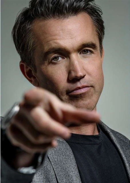 Close-up portrait of a white adult male in a grey coat and black t-shirt pointing directly at the viewer with a neutral expression.
