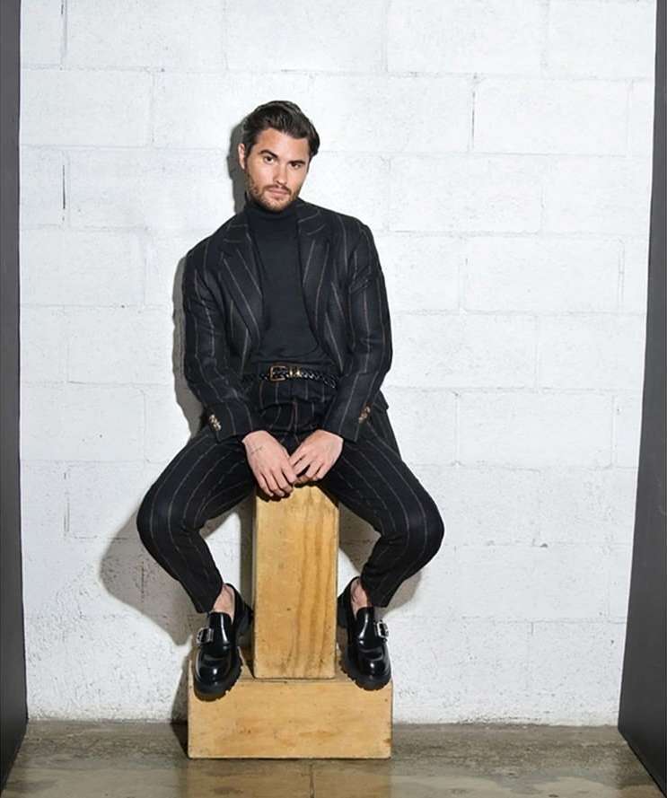 Man in pinstripe suit seated on wood block in studio. He wears a black turtleneck, leather belt, and black loafer shoes.