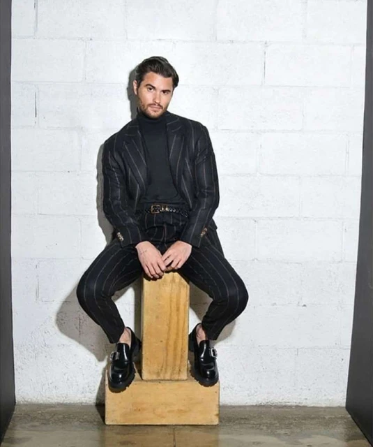 Man in pinstripe suit seated on wood block in studio. He wears a black turtleneck, leather belt, and black loafer shoes.