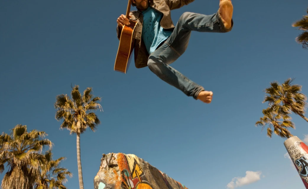 Barefoot adult male jumping in the air playing guitar with palm trees against blue sky in the background, captured at eye-level angle.