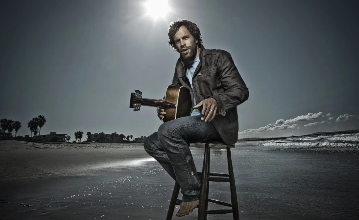 A bearded man seated on a stool on a sandy beach holding an acoustic guitar under a bright sun by photographer Sheryl Nields.