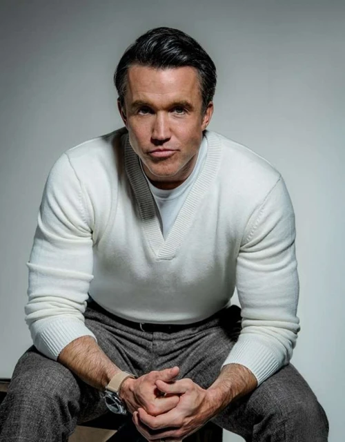 Studio shot of an adult male in a white sweater and grey trousers. He's seated with his hands clasped, against a neutral grey background.