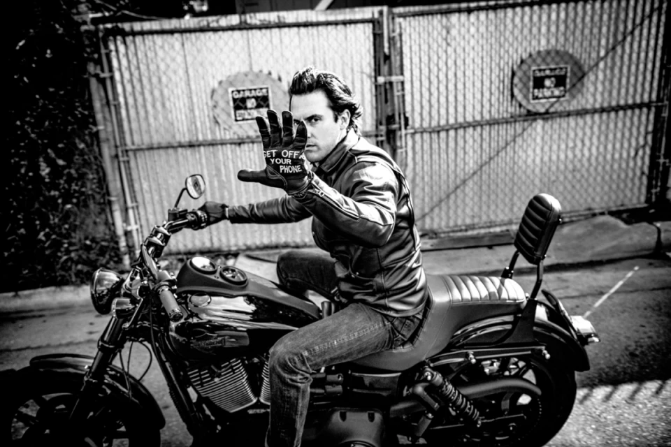 Monochrome shot of a man on a motorcycle wearing gloves that say 'Get Off Your Phone', posing in front of a metal gate.