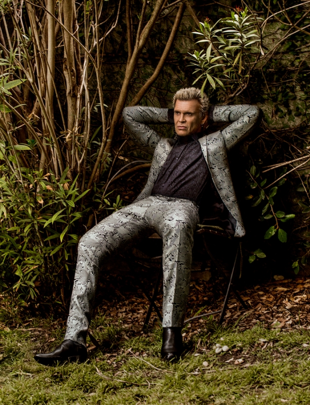 Billy Idol seated outdoors with hands behind head wearing a patterned grey suit by photographer Sheryl Nields