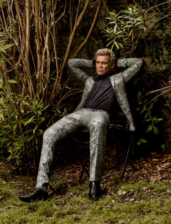 Billy Idol seated outdoors with hands behind head wearing a patterned grey suit by photographer Sheryl Nields