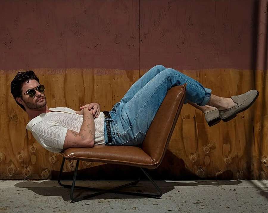 Man in casual wear, sunglasses, reclines in a leather chair against a brown wall in a relaxed pose looking to the left.