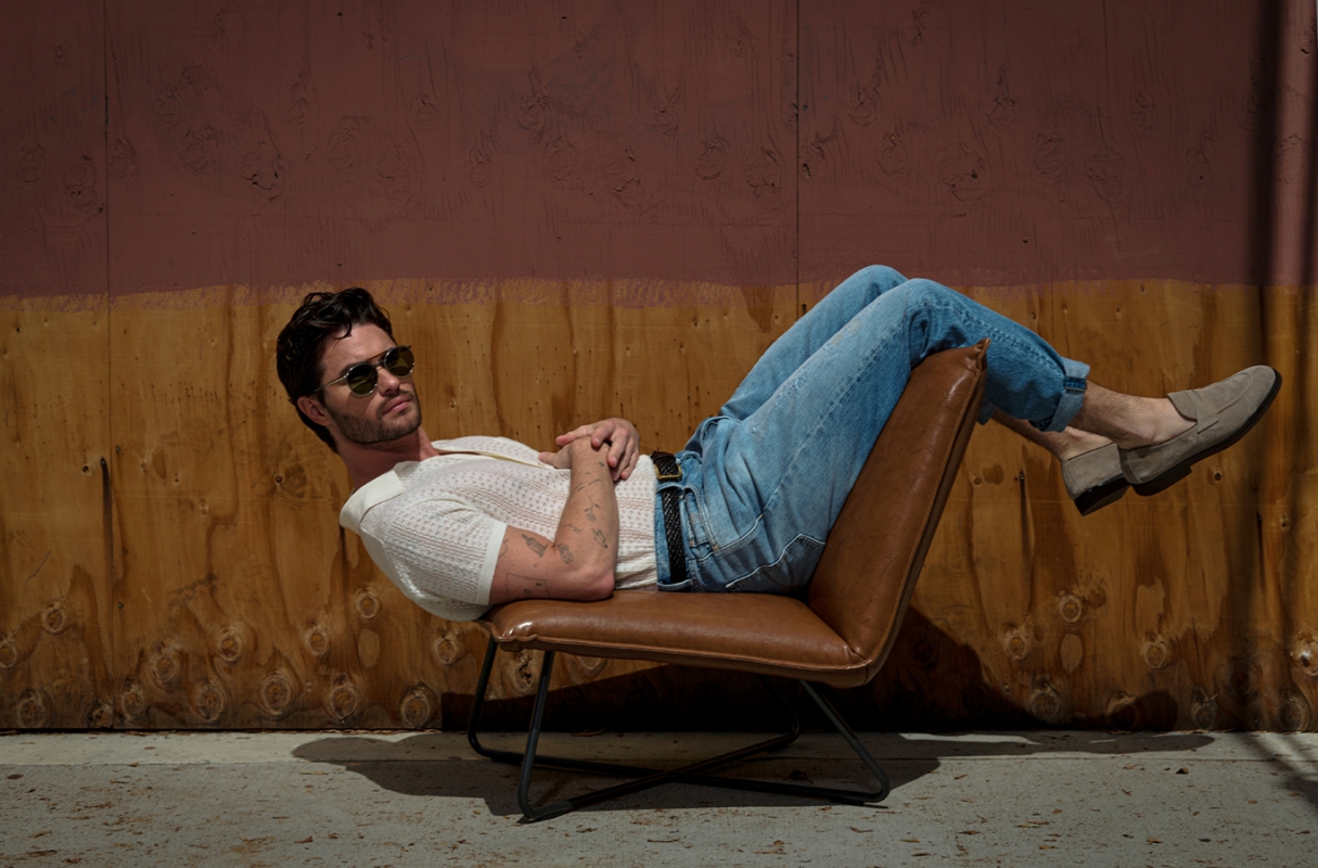 Man in sunglasses and jeans reclines on a modern chair against a textured wall in sunlight. Casual fashion portrait.