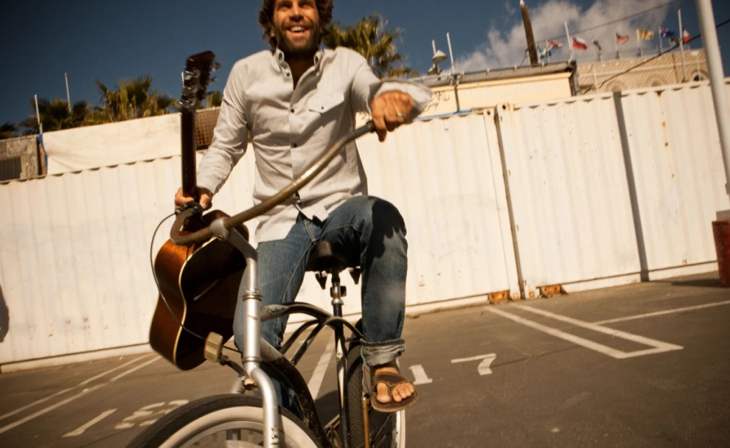 Smiling adult man riding a bicycle in an outdoor parking lot with guitar in hand under a clear sky during daytime.