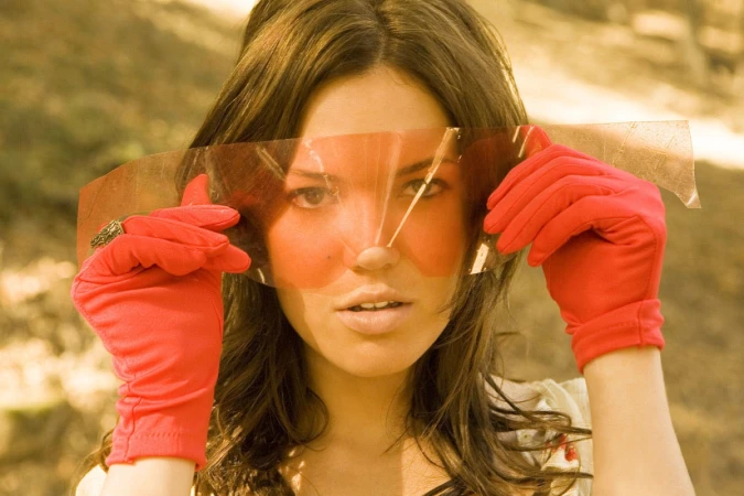 Portrait of a woman wearing bright red gloves holding a tinted translucent film strip across her eyes.