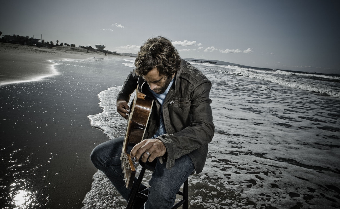 A man sits on a stool playing an acoustic guitar on a beach, waves lapping around him, with a pier visible in the distance.