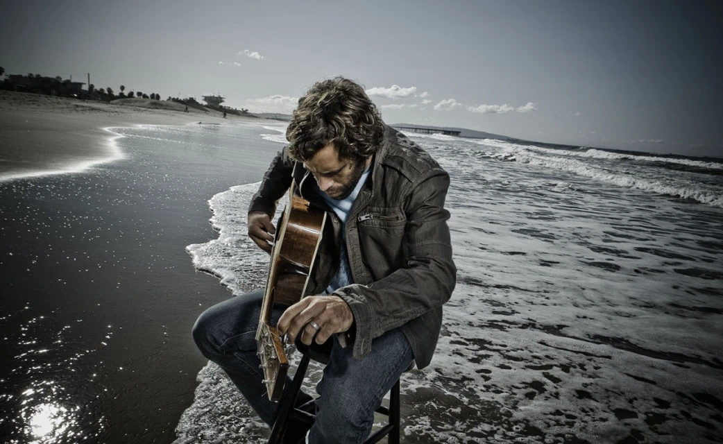 A man sits on a stool playing an acoustic guitar on a beach, waves lapping around him, with a pier visible in the distance.