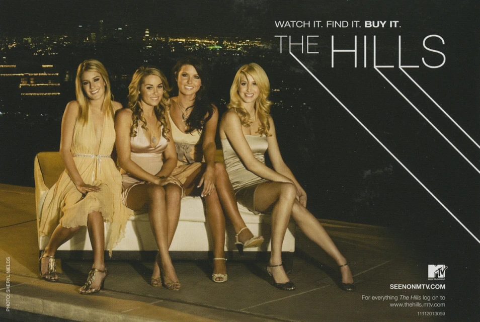 Four women sit on a white couch at night with city lights in the distance. They are wearing dresses and heels and smile at the camera.