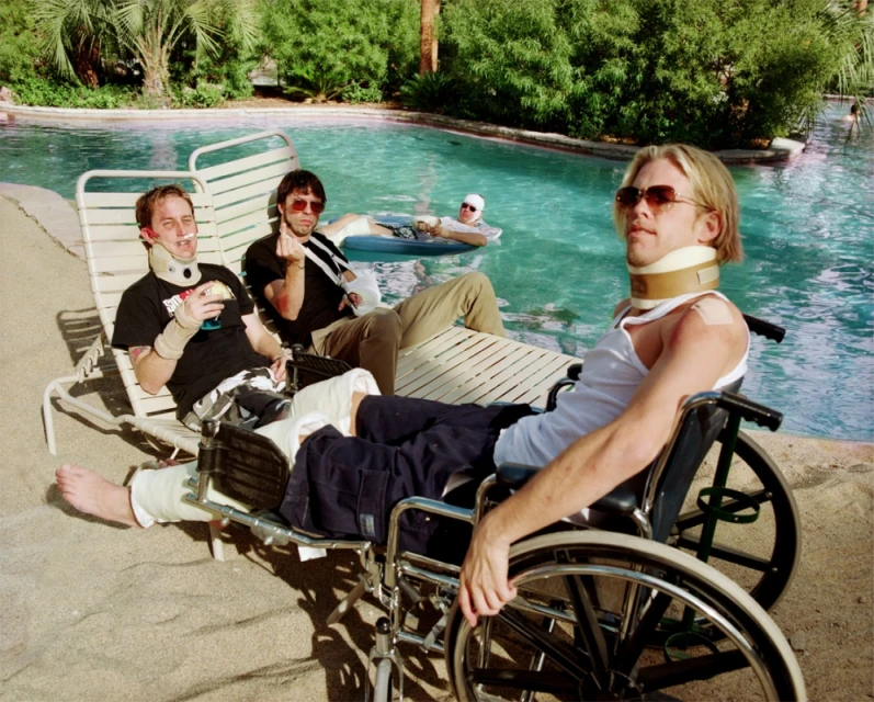 Three men with injuries and neck braces relax poolside, one in wheelchair, enjoying the resort's sunny, outdoor environment with a pool.