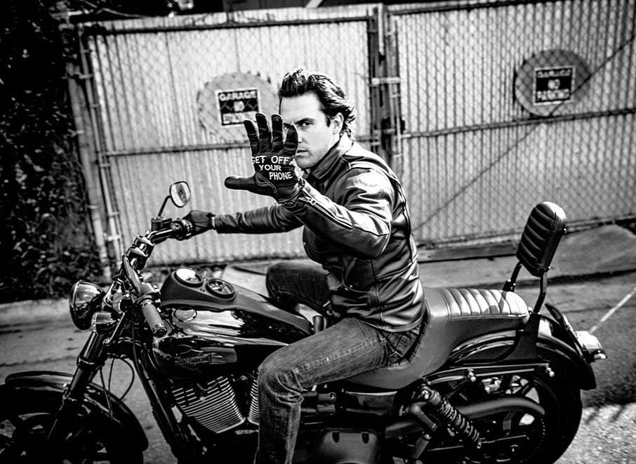 Man on a motorcycle wears a glove that reads 'Get Off Your Phone' in a black and white image, with a garage door in the background.