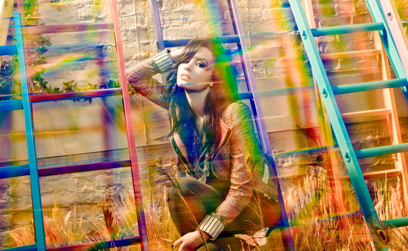 A young adult female model poses crouching next to colorful ladders near a brick wall with rainbow prism light effect and wheatgrass.