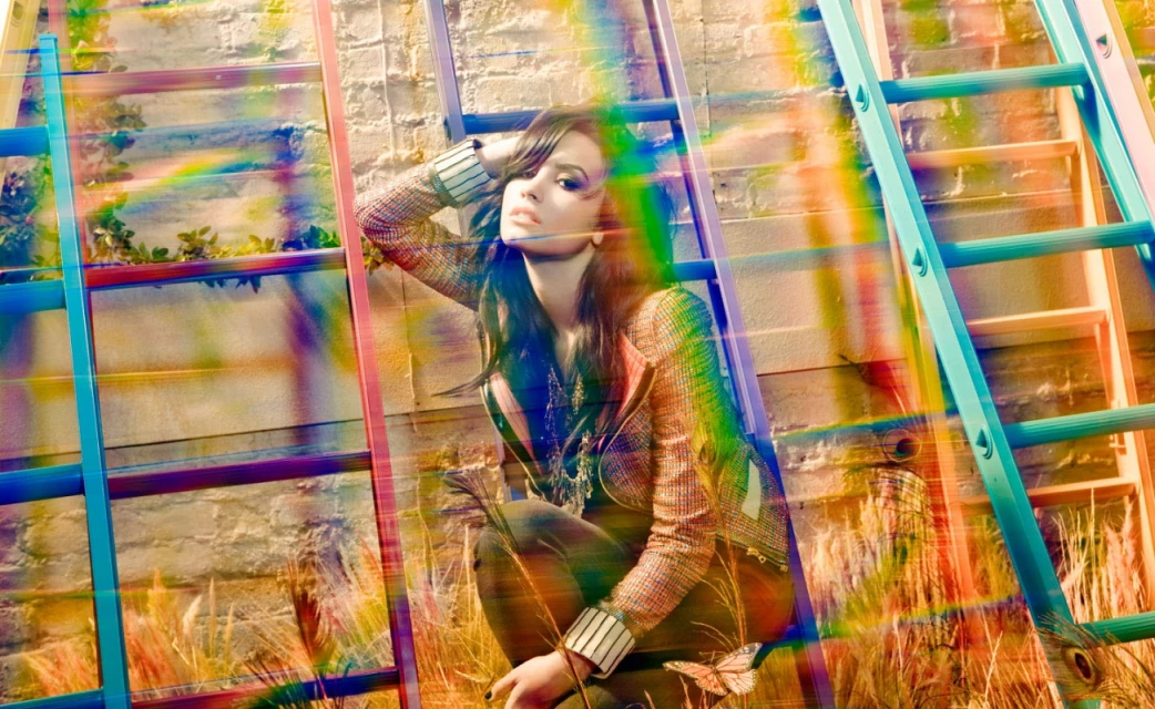 A young adult female model poses crouching next to colorful ladders near a brick wall with rainbow prism light effect and wheatgrass.