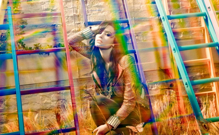 A young adult female model poses crouching next to colorful ladders near a brick wall with rainbow prism light effect and wheatgrass.