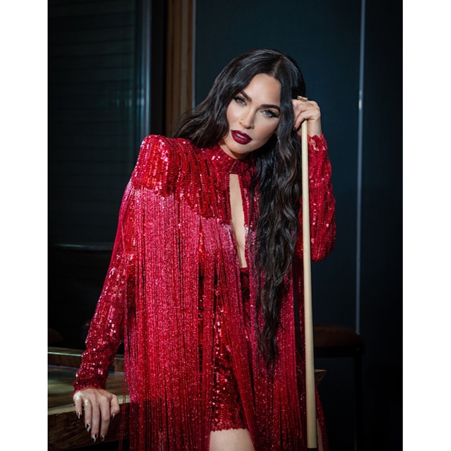 Elegant Megan Fox poses indoors in a red sequin dress and fringe jacket, holding a pool cue, with dark hair and makeup.