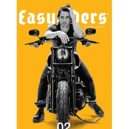Milo Ventimiglia sitting astride a black motorcycle against a solid yellow background by Sheryl Nields