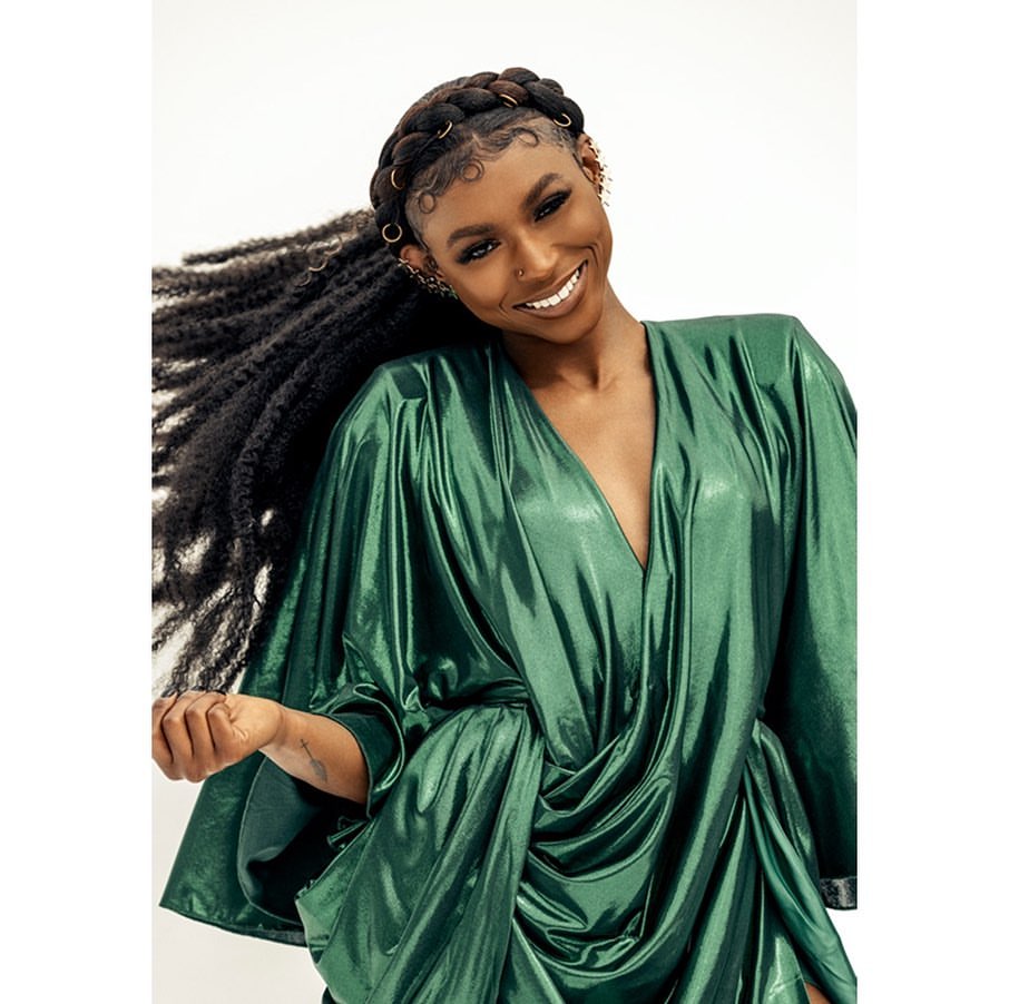 Portrait of a smiling woman with locs and a braided crown wearing a shimmery green dress against a plain white backdrop.